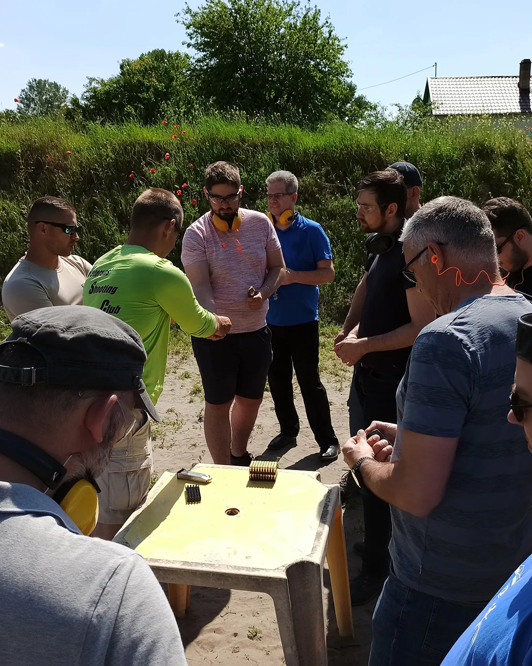 A csapat a lőtéren, fülvédővel a nyakban egy asztal körül, amelyen a tárak és a töltények sorakoznak.